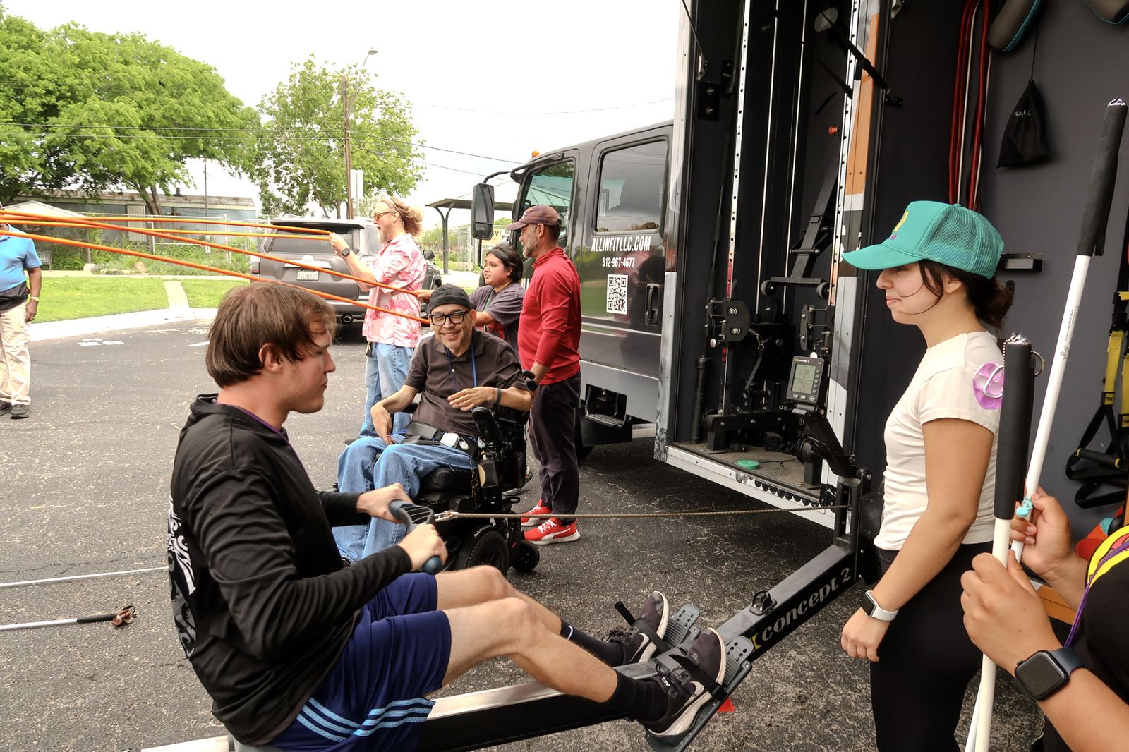 A participant using a rowing machine while a coach and others look on beside the mobile gym
